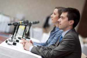 Two professionals, a man and woman, sitting at a conference table with microphones in front of them, listening attentively to Andrew Gross.
